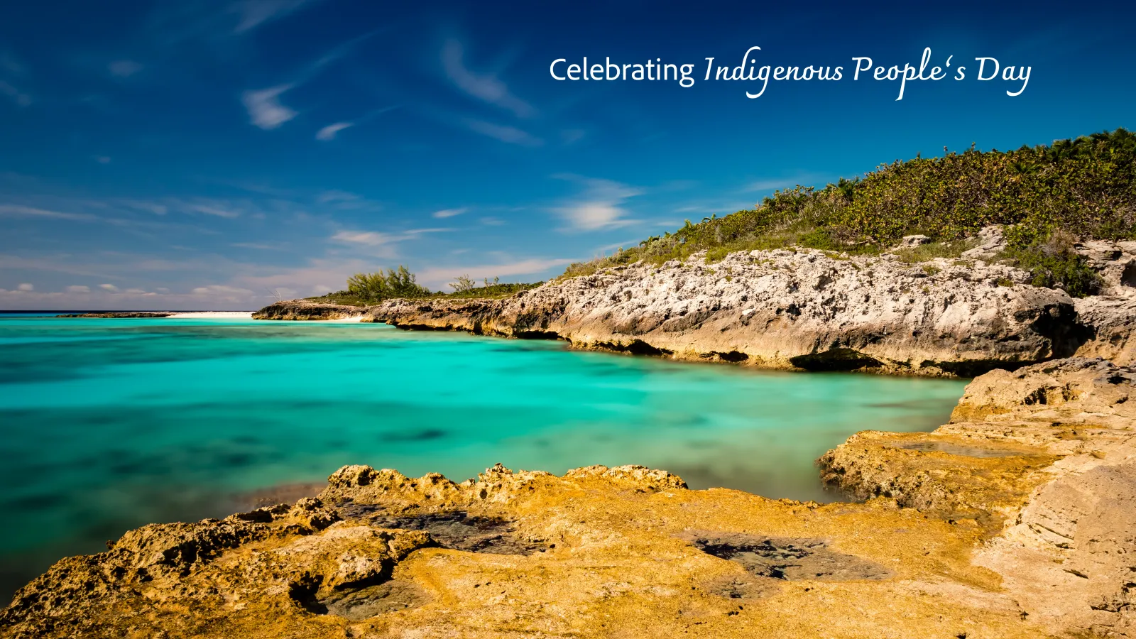 Half Moon Cay in San Salvador near Columbus' landing place. Celebrating Indigenous People's Day text in white over blue sky and water with rocky beach.