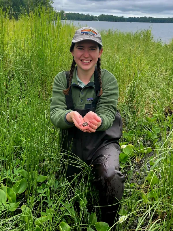 Sidney Ryan wearing chest waders and kneeling in marsh on a cloudy day