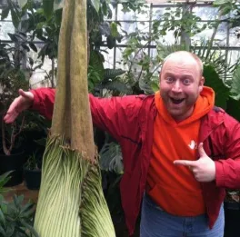 Macrander standing with right arm around spathe of Titan arum and left hand pointing to the plant